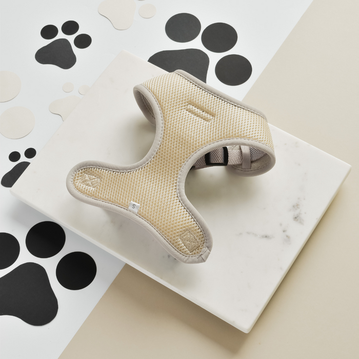 A beige dog harness from Cocopup London is displayed on a white rectangular platform, set against a background with black and beige paw print patterns. Styled in brown tweed herringbone fabric, the Brown Tweed Adjustable Neck Harness boasts a breathable mesh design, making it perfect for comfortable and lightweight dog walks.