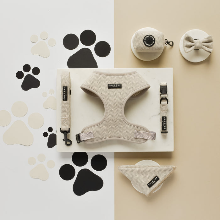 A set of Cocopup London's Brown Tweed adjustable neck harness and matching dog accessories displayed on a white and beige background with black paw print motifs. The collection includes an adjustable dog harness, leash, collar, poop bag holder, bowtie, and bandana. The accessories are sleek and well-coordinated in color and design.