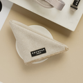 A brown tweed-patterned fabric napkin, resembling a Cocopup London Brown Tweed Bandana, folded into a triangular shape rests on a white circular dish. The napkin features a black label with white text reading "COCOPUP LONDON." Various beige accessories are visible in the background.
