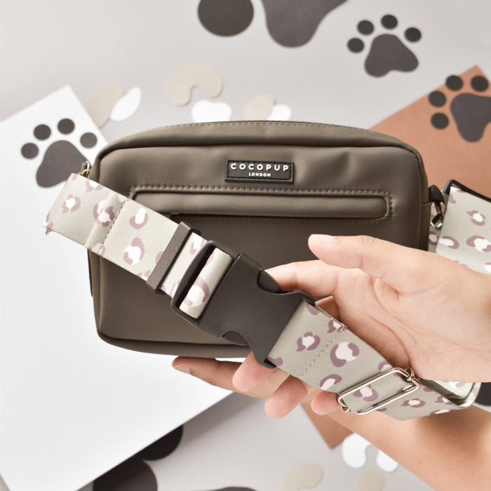 Hand holding a mocha brown Cocopup London bum bag with a patterned strap, on a surface scattered with paw print papers.