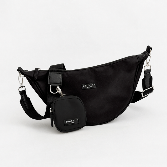 A plain black crescent shaped crossbody bag with silver metal hardware, set against a plain white background. Attached is a plain black bag strap and a small pouch for dog treats.