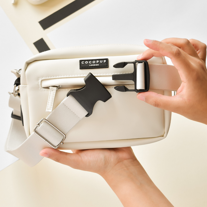 Hand adjusting the buckle on an oyster white Cocopup London bum bag with a front zip pocket.