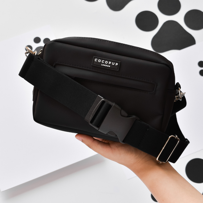 Hand holding a plain black Cocopup London bum bag with a wide strap; background features paw print pattern.