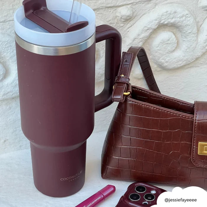 Burgundy stainless steel tumbler by Cocopup London next to a purse, lipstick, and car keys on a light surface.