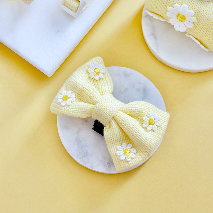 Yellow knitted dog bow tie with white daisy pattern, shown on a marble dish.