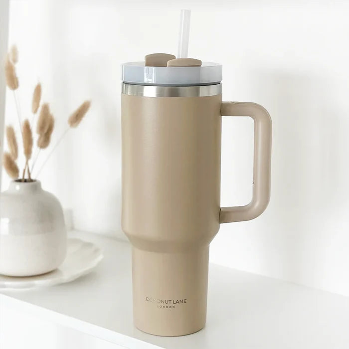 Caramel-coloured stainless steel tumbler with handle and straw by Cocopup London, set next to dried grass.