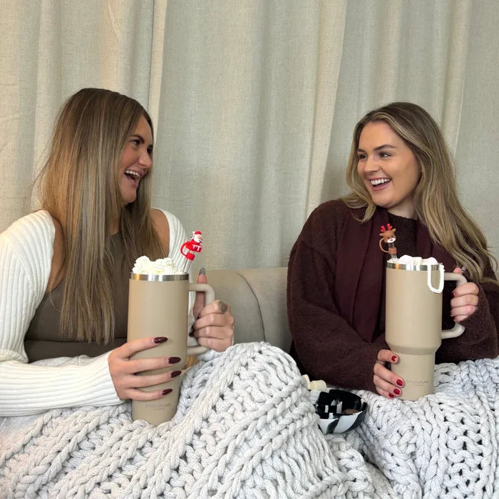 Two women on a couch with mugs and a beige stainless steel Cocopup tumbler, wrapped together in a chunky knit blanket.