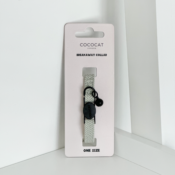 Sage green tweed cat collar with breakaway buckle and black bell, shown on a white shelf.