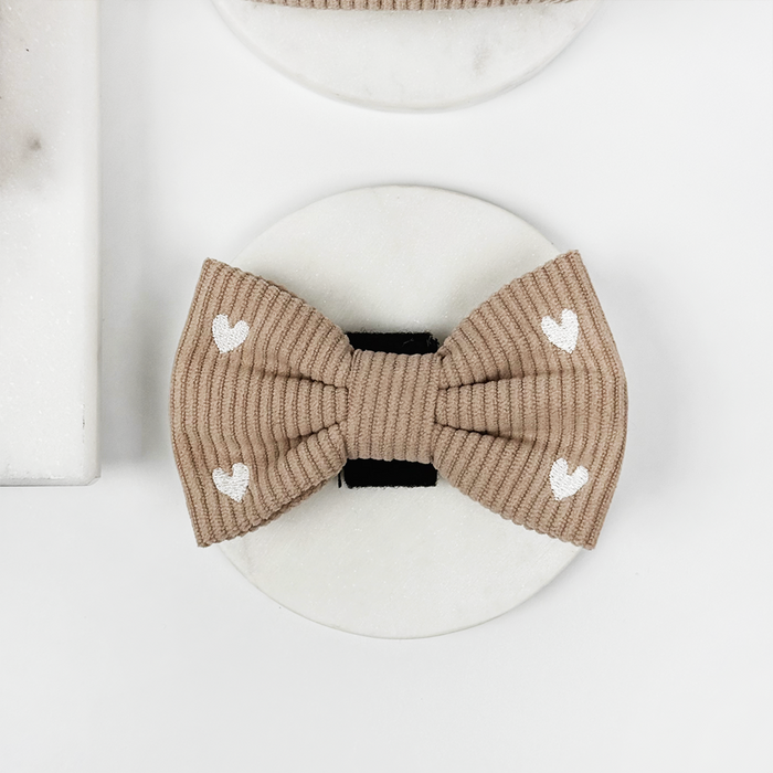 Tan ribbed dog bow tie with small white hearts, displayed on a plain round white surface.