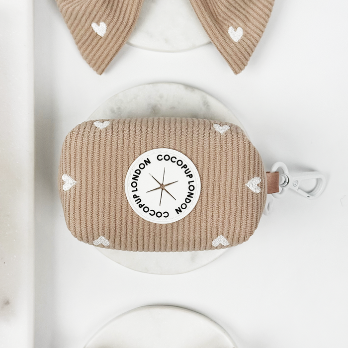 Beige ribbed poop bag holder with white hearts and metal clip, by Cocopup London, on a white marble background.