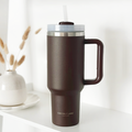 Chocolate stainless steel tumbler with handle and straw by Cocopup London on a shelf beside dried flowers.