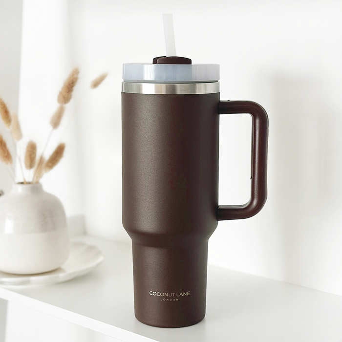 Chocolate stainless steel tumbler with handle and straw by Cocopup London on a shelf beside dried flowers.