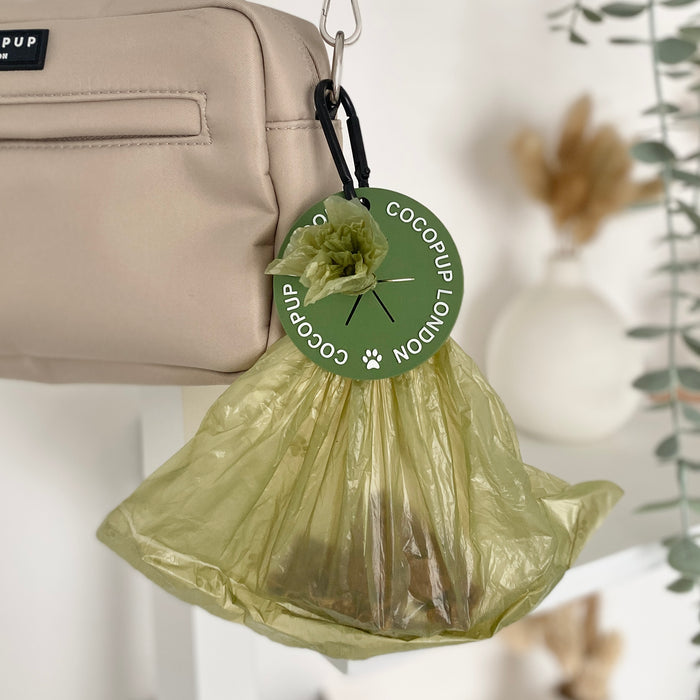 Khaki CocoPOOP Pocket attached to a beige bag, shown among green plants.
