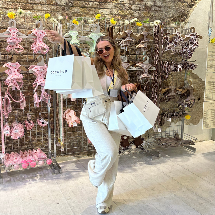 Woman with shopping bags stands by a metal rack displaying Cocopup London dog accessories in gift bags.