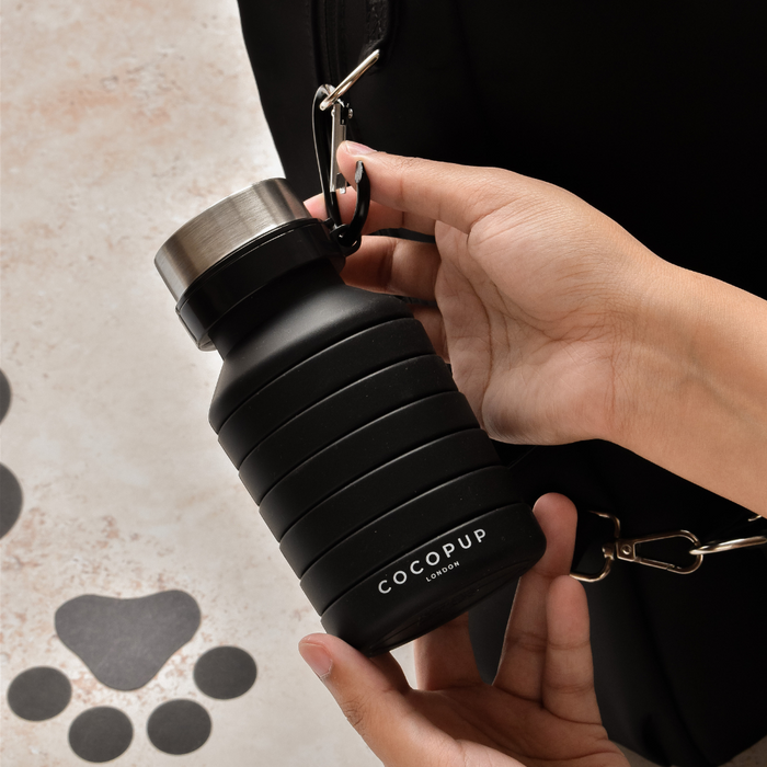 Hand attaching a black Cocopup London collapsible water bottle to a bag beside paw print patterns on the floor.
