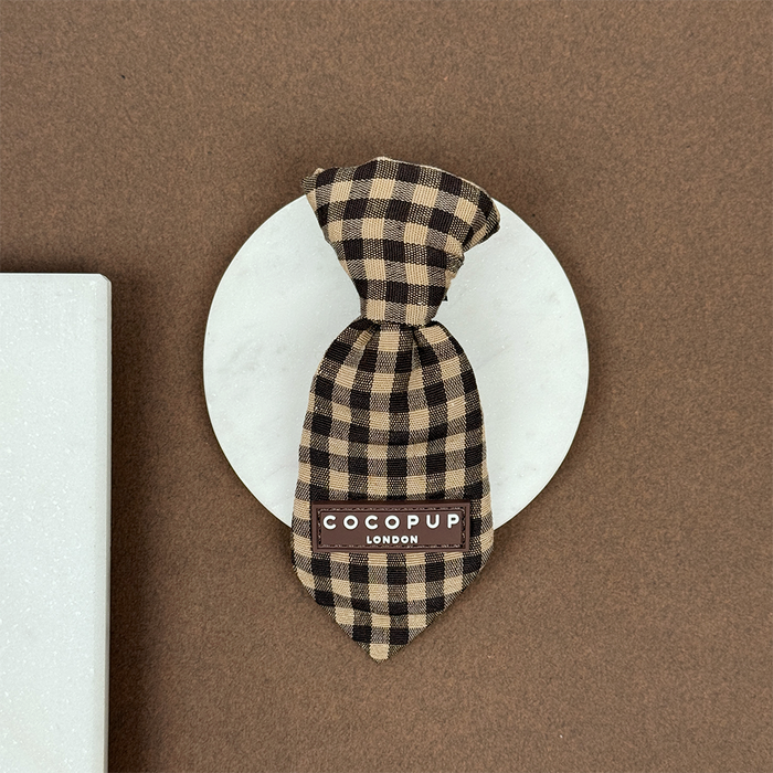 Brown and beige check dog tie from Cocopup London displayed on a round white surface.