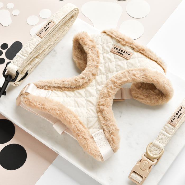 Discover the Cocopup London Quilted Adjustable Neck Harness set, designed with stylish quilted cream fabric and faux fur trim. This plush harness features a fully adjustable chest strap for the perfect fit. The set includes a matching cream-colored leash and collar, both adorned with metal hardware. All items are elegantly displayed on a white marble surface featuring black and beige circles.
