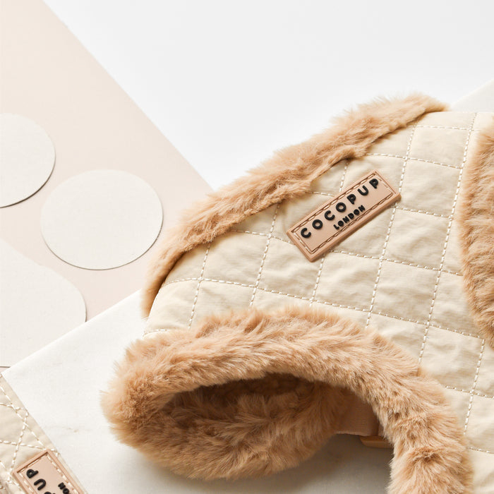 A close-up of the stylish beige quilted Adjustable Neck Harness with faux fur trim. A label reading "Cocopup London" is visible on the harness. Circular paper cutouts are scattered beside the fully adjustable chest strap on a white and beige surface.