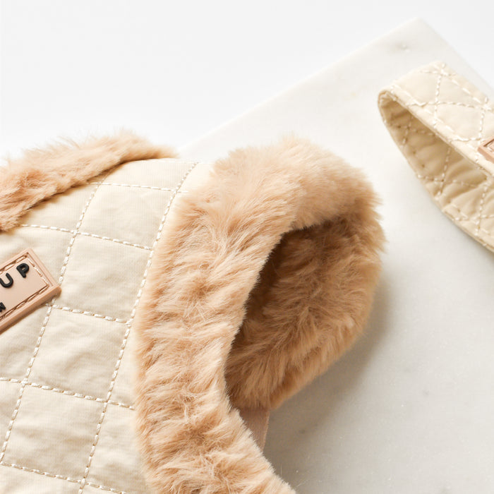 A close-up image of a luxurious pet accessory. The stylish Cocopup London Quilted Adjustable Neck Harness is quilted and features a soft, tan fur lining. An embossed tag with the text "PUP" is visible on the fabric. Another strap of the same material is partially seen on the right.