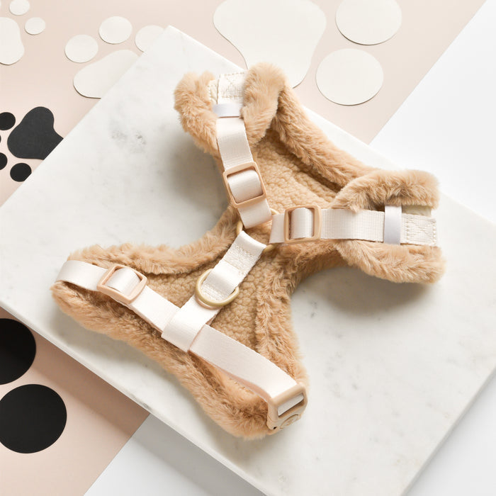 The Quilted Adjustable Neck Harness by Cocopup London is a stylish, fuzzy beige dog accessory with adjustable white straps and clips. Displayed on a white marble slab, it's surrounded by abstract circular cutouts in black, beige, and white. This harness boasts a plush texture and fully adjustable chest strap.