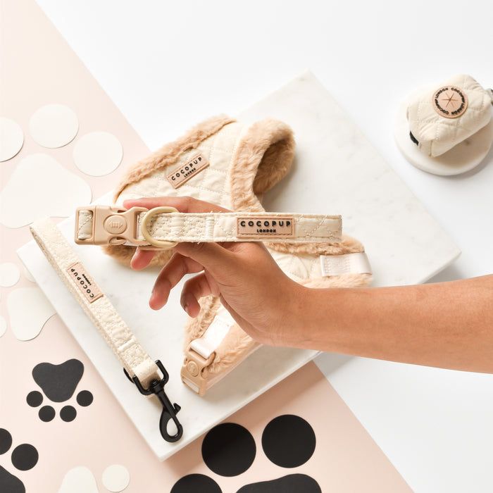 Hand holding cream quilted collar by Cocopup London beside matching leash and harness on marble tray with paw prints.