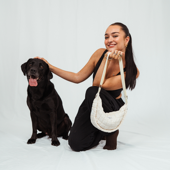 Woman kneeling next to black dog, holding a light beige crescent-shaped Cocopup crossbody dog walking bag.