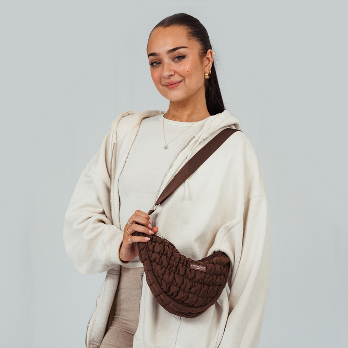 Smiling woman in cream hoodie wears a brown Cocopup London crossbody crescent bag.