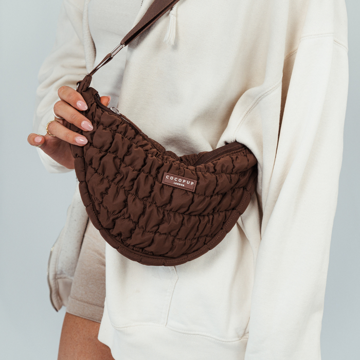 Person in a cream hoodie holds a brown Cocopup London crossbody crescent dog walking bag.