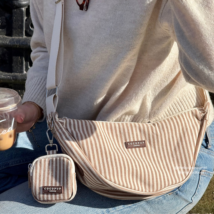 Person in cream sweater holding iced coffee and a striped Cocopup London crossbody dog walking bag.