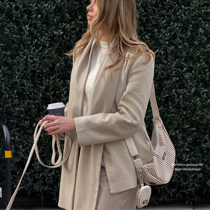 Woman in beige outfit holds a coffee cup and striped linen Cocopup London crossbody dog walking bag.