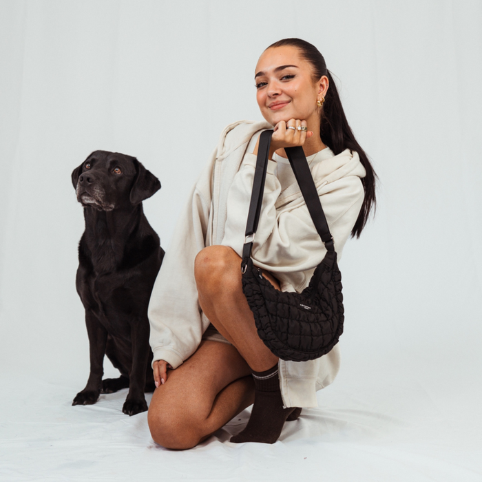 Woman kneeling next to her black dog with a black Cocopup London crescent crossbody dog walking bag at her side.