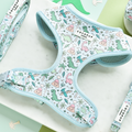 A pastel blue Dinopaws Adjustable Neck Harness with a whimsical dinosaur print featuring green, pink, and blue dinosaurs alongside tropical trees and plants. The adjustable neck harness includes the brand name "Cocopup London" on a small label. Displayed on a light marble surface with matching accessories.