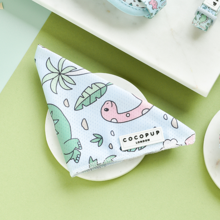 A triangular, light blue Bandana - Dinopaws from Cocopup London, adorned with a playful dinosaur and leaf pattern, lies flat on a white marble surface. The bandana features a small white label that reads "COCOPUP LONDON" near the bottom edge. The background showcases a light green surface, accentuating this charming piece from the Cocopup range.