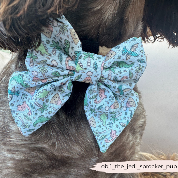 Curly brown dog wearing a Cocopup London Dinopaws Sailor Bow Tie with green dinosaurs and palm trees.