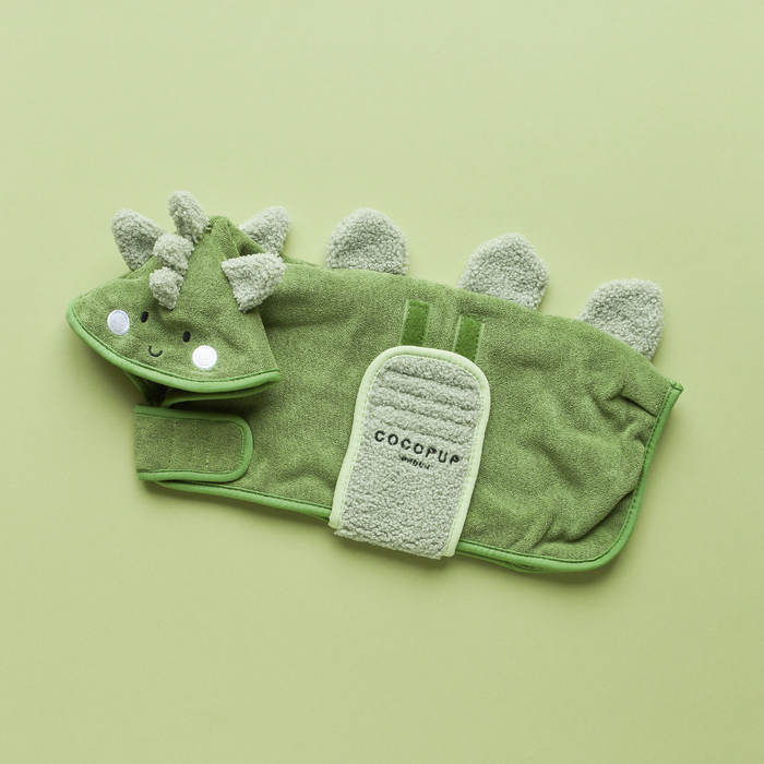 A green PRE-ORDER Dinopup Drying Robe from Cocopup London is laid out on a light green background. The robe features a hood with an adorable dinosaur face, fabric spikes along the back, and a front pocket labeled "COCCUP." It boasts Premium Microfibre material and lighter green trim.