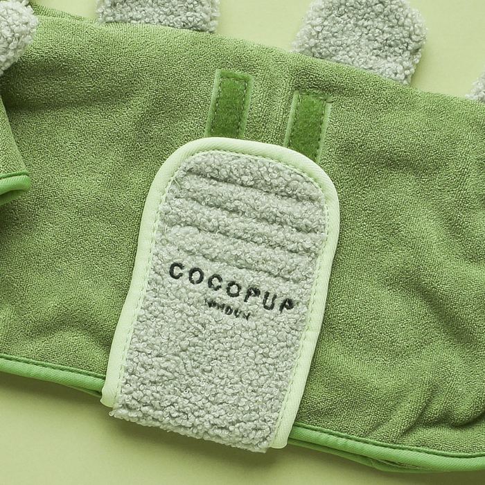 A green PRE-ORDER Dinopup Drying Robe from Cocopup London, featuring a soft, textured, and fuzzy appearance, is displayed against a matching green background. Made from premium microfibre, the robe has a rectangular stitched area with the brand name "Cocopup" and the word "London" written beneath it.