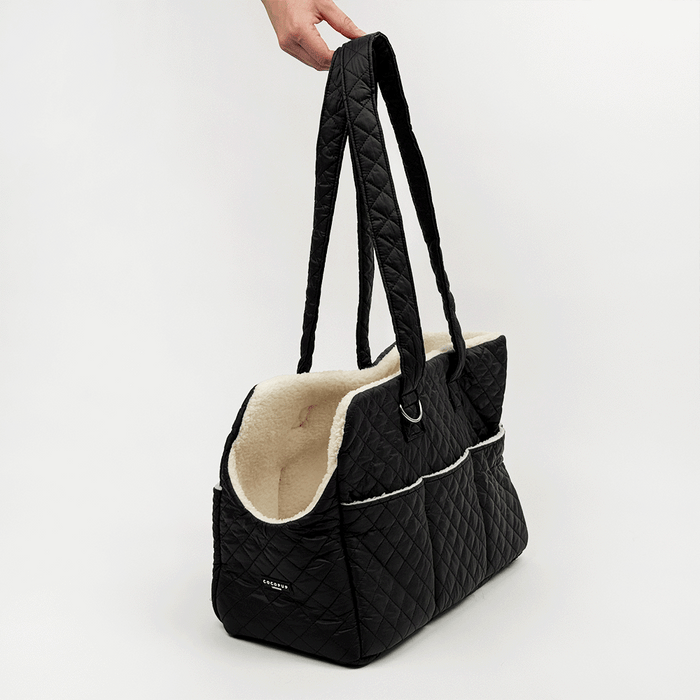 Black quilted Dog Carrier with a cream sherpa fleece lining, featuring a diamond-quilted exterior, a side pocket with white piping, and a small silver-tone metal ring detail.
