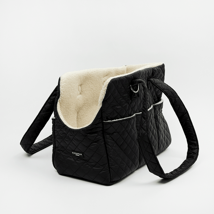 Black quilted Dog Carrier with a cream sherpa fleece lining, featuring a diamond-quilted exterior, a side pocket with white piping, and a small silver-tone metal ring detail.