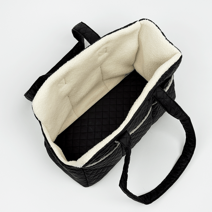 Showing the inside of a black quilted Dog Carrier with a cream sherpa fleece lining, featuring a diamond-quilted exterior, a side pocket with white piping, and a small silver-tone metal ring detail.