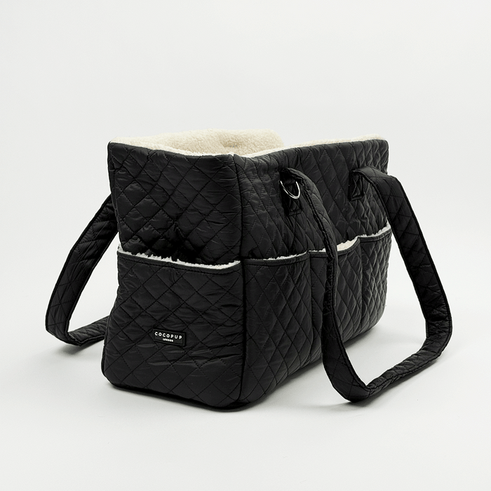 Showing the back of a black quilted Dog Carrier with a cream sherpa fleece lining, featuring a diamond-quilted exterior, a side pocket with white piping, and a small silver-tone metal ring detail.