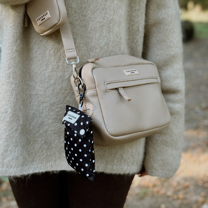 Person in beige sweater with beige crossbody bag and a dog wearing a black raincoat with white polka dots.