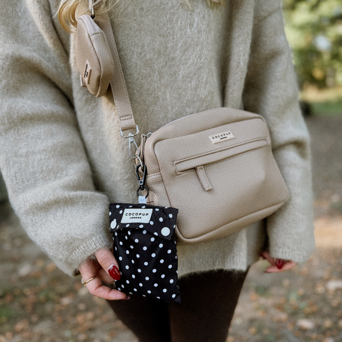 Person in beige sweater holding a black polka dot dog raincoat and matching pouch from Cocopup London outdoors.