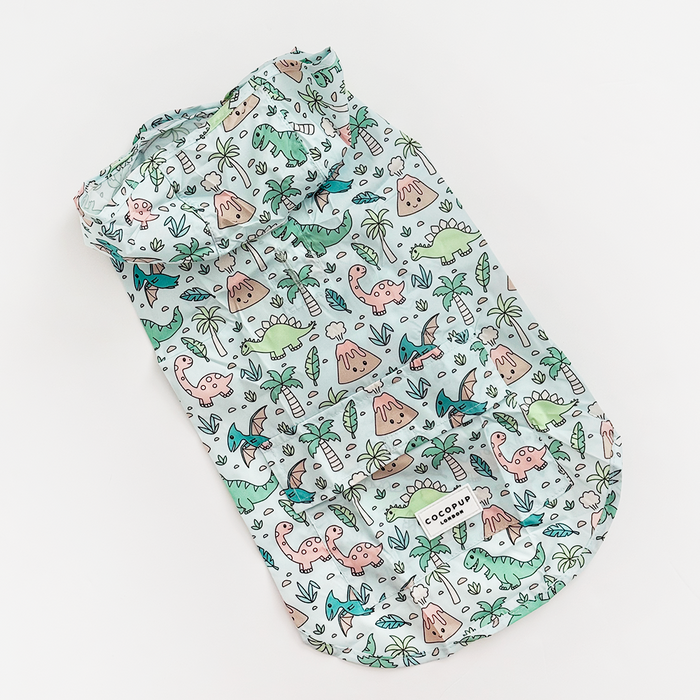 Light blue Cocopup London dog raincoat with dinosaur, palm tree, and volcano print, displayed on a white background.