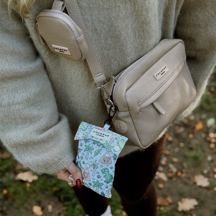 Person in beige sweater holds Dinopaws-print dog raincoat and waste bag, wearing tan crossbody bags.