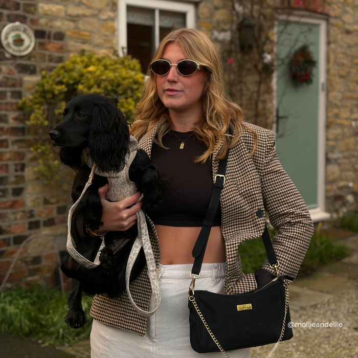 Woman in sunglasses holding a black Cocopup London Dog Walking Bag with her black dog outside a brick house.