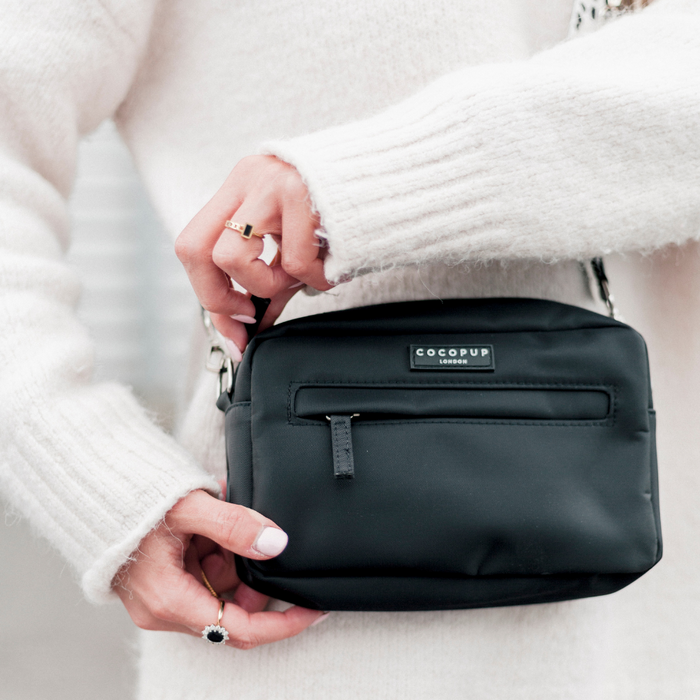 Black Cocopup London Dog Walking Bag with front zip and poop bag dispenser held by woman in a white knit sweater.