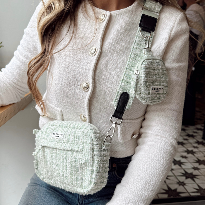 Woman in a white cardigan holding two green textured tweed Cocopup London bag straps at a wooden table.