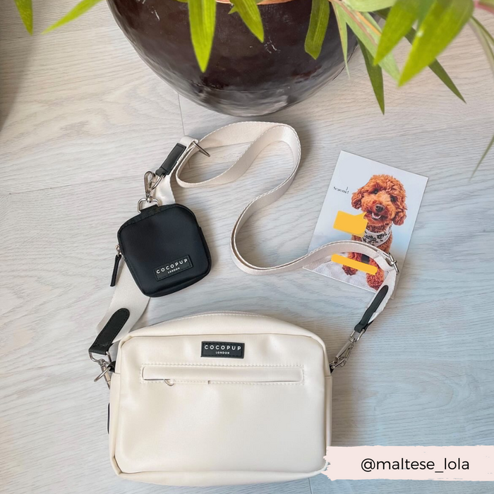 Oyster white Cocopup London dog walking bag with black poop bag dispenser on a light floor, next to a postcard and plant pot.