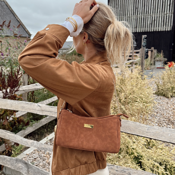 Blonde woman with a tan suedette Cocopup London dog walking bag near a wooden fence outdoors.
