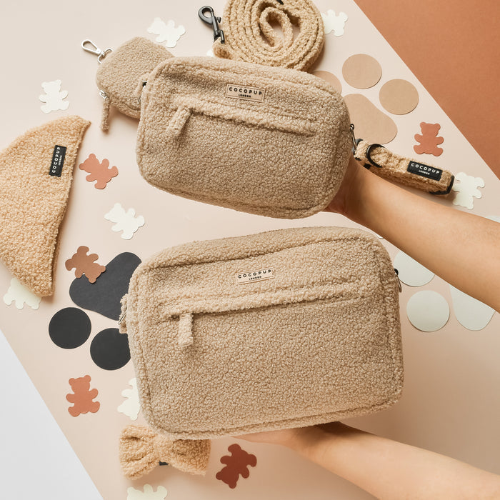 Two hands hold a plush, tan Cocopup London Dog Walking Bag; a teddy bear and abstract shapes are in the background.